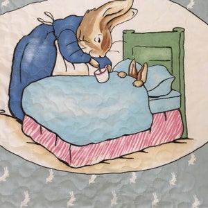 May include: A blue and brown rabbit in a blue dress gives a cup of tea to a brown rabbit in bed. The bed has a green headboard and a pink and white striped bed skirt. The background is a light blue with white rabbit silhouettes.
