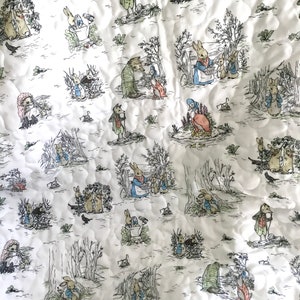 May include: A white fabric quilt with a repeating pattern of Peter Rabbit characters in black and white. The characters include Peter Rabbit, Mr. McGregor, and other animals from the story.