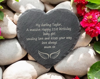May include: A heart-shaped slate stone with engraved text: "My darling Taylor, A massive Happy 21st birthday baby girl sending love and kisses your way love always mum xx" and a wing design. The stone is surrounded by white stones and red flowers.