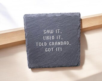 May include: A square, dark gray slate coaster with the text "SAW IT, LIKED IT, TOLD GRANDAD, GOT IT!" in white lettering. The coaster is placed on a light-colored wooden surface.