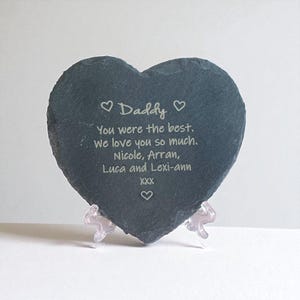 May include: A heart-shaped slate memorial with the word "Daddy" and a loving message engraved in white. The message reads "You were the best. We love you so much. Nicole, Arran, Luca and Lexi-ann XXX". It rests on a clear plastic stand.