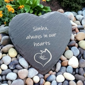 May include: A heart-shaped slate stone memorial with the inscription "Simba, always in our hearts" and a small cat silhouette. The stone is surrounded by small, colorful pebbles.