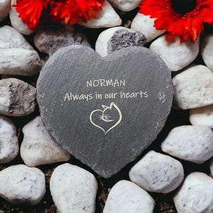 May include: A gray heart-shaped slate stone with the inscription "NORMAN Always in our hearts" and a small engraved image of a cat inside a heart. The stone is surrounded by white pebbles and red flowers.