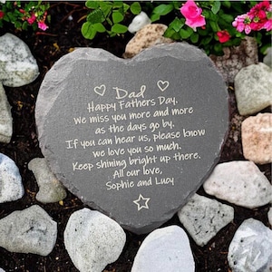 May include: A heart-shaped slate memorial stone with engraved text. The inscription reads "Dad Happy Fathers Day. We miss you... Keep shining bright up there. All our love, Sophie and Lucy." The stone is set amongst rocks and flowers.