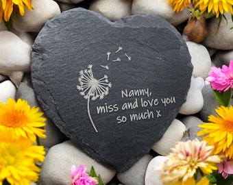 May include: A heart-shaped, dark gray slate stone with the engraved text "Nanny, miss and love you so much x" and a dandelion design. The stone is surrounded by gray stones, yellow and pink flowers.