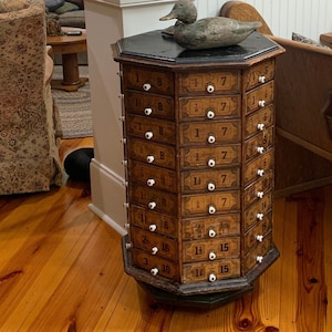 May include: An antique wooden cabinet with multiple drawers, each labeled with numbers and white knobs. A black lamp with a black shade sits atop the cabinet, and a wooden duck figurine rests on the cabinet. An antique telephone is mounted on the wall.