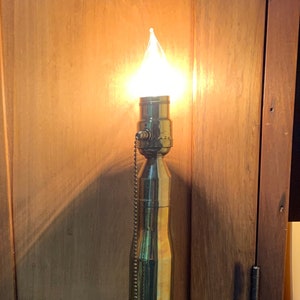 May include: A brass lamp with a pull chain switch, made from a repurposed bullet casing. The lamp is lit and casting a warm glow. The lamp is sitting on a wooden surface.