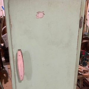 May include: A vintage green cabinet with a single door and a pink handle. The cabinet has a distressed finish and is sitting on a wooden floor.