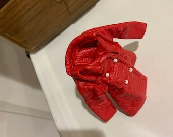 May include: A red faux leather jacket with white button accents. The jacket is folded and lying on a white surface.