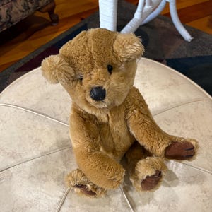 May include: A plush teddy bear with brown fur, a black nose, and brown paws. The bear is sitting on a cream-colored, round ottoman. The bear's ears are rounded and the fur is soft and fluffy.