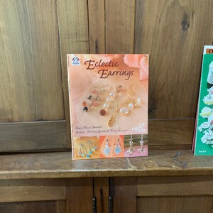 May include: A book titled "Eclectic Earrings" with a pink and white floral background. The book cover features a variety of earrings with pearls, beads, and other decorative elements. The text "Over a Dozen Beautiful Projects. Fabulous Accents for Every Occasion." is printed on the cover.