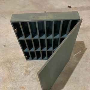 May include: A teal blue wooden cabinet with a hinged door and multiple compartments. The cabinet has a brass knob and a brass hook on the top.