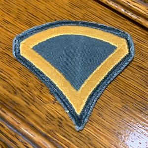 May include: A dark blue military patch with a yellow chevron design. The patch is made of fabric and has a stitched border.