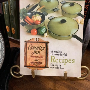 May include: A cookbook titled "Country Inn Cookware by West Bend" with a picture of a green pot with vegetables and three other green pots on a counter. The text "A wealth of wonderful Recipes for every occasion..." is printed on the cover.