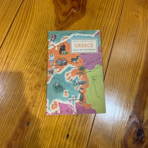 May include: A vintage travel guide titled "Greece" from the "Around the World Program" series. The cover features a colorful map of Greece with illustrations of people and landmarks. The book is set against a wooden background.