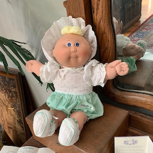 May include: A Cabbage Patch Kid doll with blonde hair, blue eyes, and a green and white dress. The doll is sitting on a wooden surface with a book and a piece of paper.