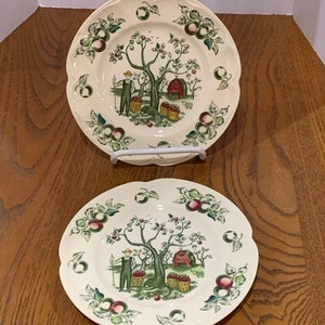 May include: Two white ceramic plates with a green, red, and brown apple orchard design. The plates feature a man picking apples from a tree and a small red barn.
