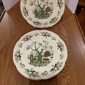 May include: Set of three white ceramic dinner plates with a green, red, and brown apple orchard design. The plates feature a stylized apple tree with red apples, a red barn, and a person picking apples.