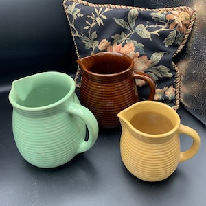 May include: Three ceramic pitchers in different colors and sizes. The largest pitcher is brown with a ribbed design. The medium pitcher is green with a ribbed design. The smallest pitcher is yellow with a ribbed design.