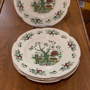 May include: A set of four white ceramic dinner plates with a scalloped edge. Each plate features a green and red apple tree with a red barn and a person picking apples. The plates are decorated with green leaves and red apples.