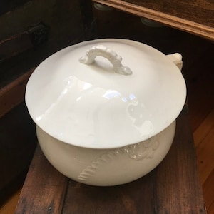 May include: White ceramic chamber pot with a lid and a handle. The pot has a decorative design on the side. The pot is sitting on a wooden surface.