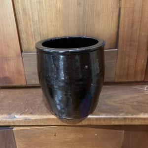 May include: A dark brown, glazed ceramic pot with a rounded body and a wide, open top. The pot has a glossy finish and appears to be empty. The pot is sitting on a wooden surface.