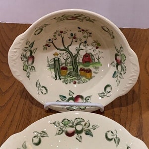 May include: Two white ceramic bowls with a green and brown apple orchard design. The bowls have a scalloped edge and a handle on each side.