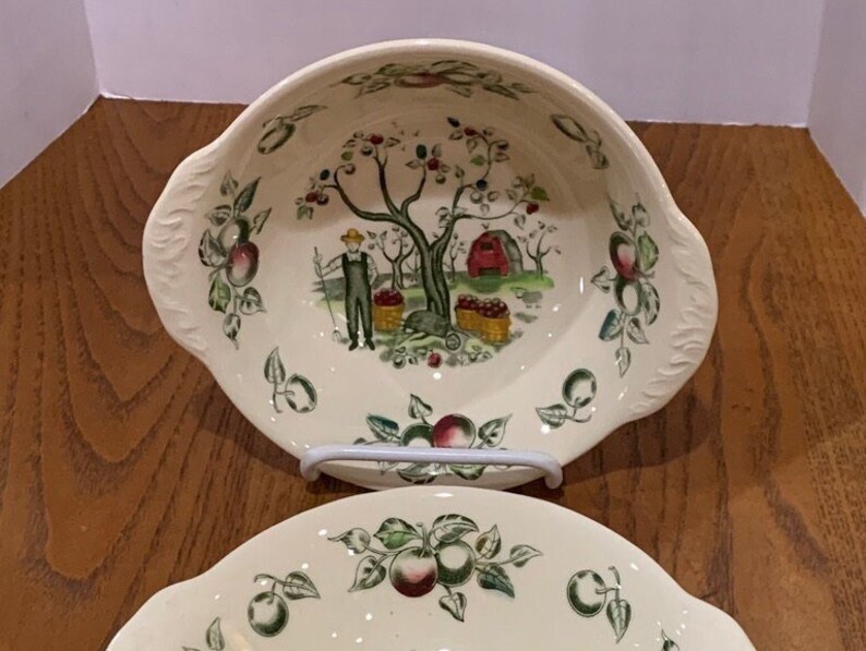 May include: Two white ceramic bowls with a green and brown apple orchard design. The bowls have a scalloped edge and a handle on each side.