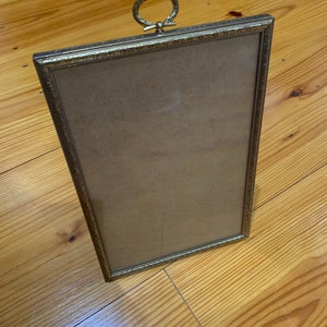 May include: A gold-toned picture frame with an ornate design and a decorative ring handle at the top.