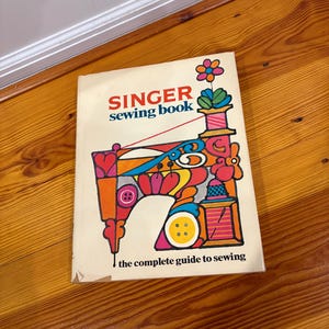 May include: A vintage Singer sewing book with a colorful illustration of a sewing machine on the cover. The cover features the text "SINGER sewing book" and "the complete guide to sewing". The book is lying on a wooden floor.