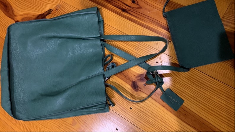 Puede incluir: Un conjunto de bolso tote y bandolera verde oscuro. El bolso tote tiene asas largas y un compartimento principal grande. El bolso bandolera tiene una correa para el hombro y forma rectangular. Ambos bolsos tienen un aspecto texturizado y una etiqueta que dice "for people".