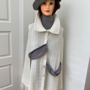 May include: A white knitted poncho with fringe trim and a button closure. The poncho is worn over a gray turtleneck and a gray scarf. The model is wearing a black and white hat.