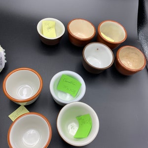 May include: A collection of eight small ceramic bowls with brown rims. The bowls are arranged in a circle on a dark surface. Some of the bowls have handwritten notes on them, including "Hell", "Franklin 3 3/8 x 2 3/8", "Jackson", and "Hall 3 1/4 x 2 3/4".
