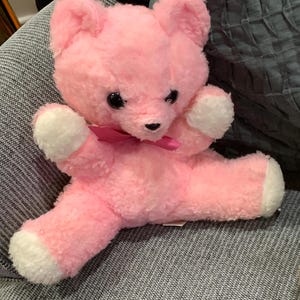 May include: A pink plush teddy bear with white paws and a pink bow. The bear is sitting on a gray textured surface.