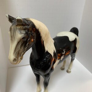 May include: A vintage plastic horse figurine in a standing pose. The horse has a black and white coat with a cream-colored mane and tail. The figurine has detailed features, including a bridle and saddle design.