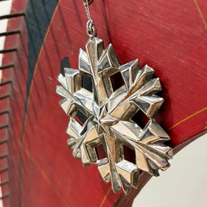 May include: A silver snowflake ornament with a detailed, geometric design hangs from a delicate chain. The ornament is set against a red background with black and gold accents, suggesting a festive or holiday theme.