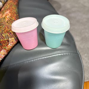 May include: Two small, colorful plastic cups with lids. One is pink, the other is teal. Both have white lids. The cups are sitting on a dark gray surface. The cups appear to be for storing small items or for portion control.