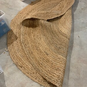 May include: A round, natural fiber rug with a woven texture. The rug is beige and has a slightly frayed edge.