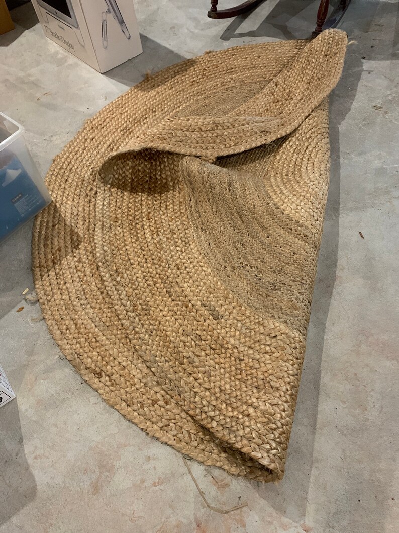 May include: A round, natural fiber rug with a woven texture. The rug is beige and has a slightly frayed edge.