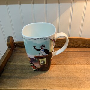 May include: A white ceramic mug with a square shape and a handle. The mug features a colorful illustration of cats in a cartoon style. The illustration includes a cat wearing a red and white striped shirt and a cat wearing a blue and white striped shirt. The mug is sitting on a wooden surface.