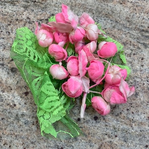 May include: A cluster of pink fabric flowers with green lace trim. The flowers are arranged in a bouquet shape and have a soft, velvety texture.