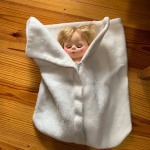 May include: A white plush baby doll sleeping bag with a zipper closure and a button detail. The doll is inside the sleeping bag and has blonde hair and blue eyes.