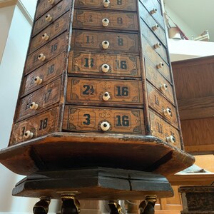 Puede incluir: Antiguo mueble de boticario de madera con numerosos cajones. El mueble es de m&uacute;ltiples lados, cada lado con m&uacute;ltiples cajones con pomos blancos y numeraci&oacute;n negra. El mueble se asienta sobre una base con ruedas para la movilidad. La madera tiene un acabado envejecido.