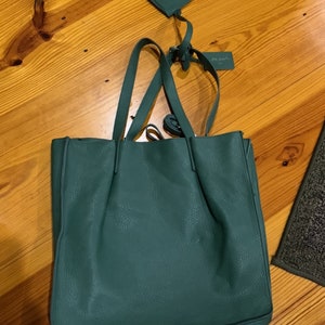 Puede incluir: Un bolso tote verde azulado con una peque&ntilde;a bolsa a juego. El bolso tote tiene asas largas y una superficie texturizada. La bolsa tiene cierre de cremallera y una correa. Ambos art&iacute;culos est&aacute;n sobre una superficie de madera.