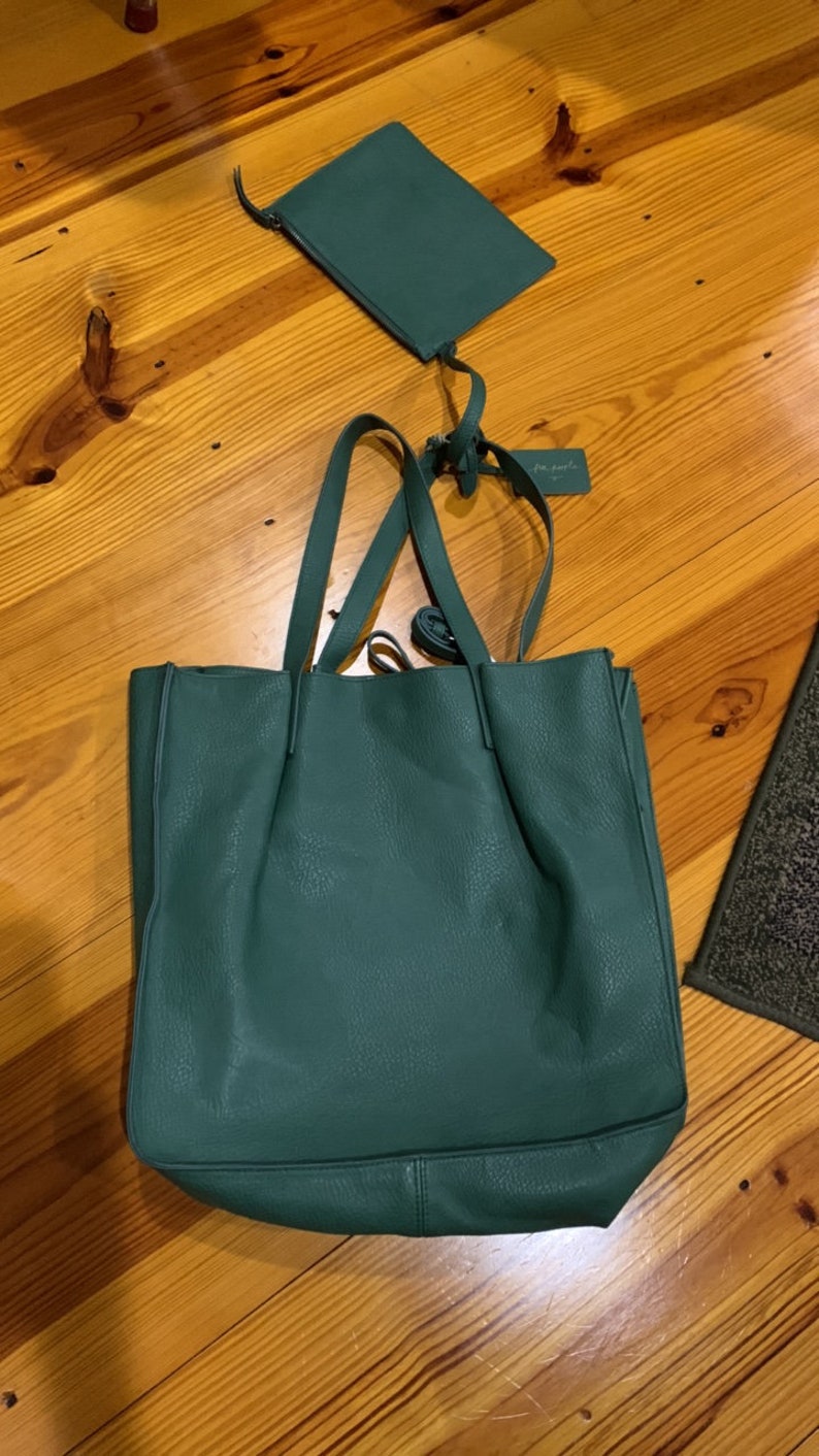 Puede incluir: Un bolso tote verde azulado con una peque&ntilde;a bolsa a juego. El bolso tote tiene asas largas y una superficie texturizada. La bolsa tiene cierre de cremallera y una correa. Ambos art&iacute;culos est&aacute;n sobre una superficie de madera.