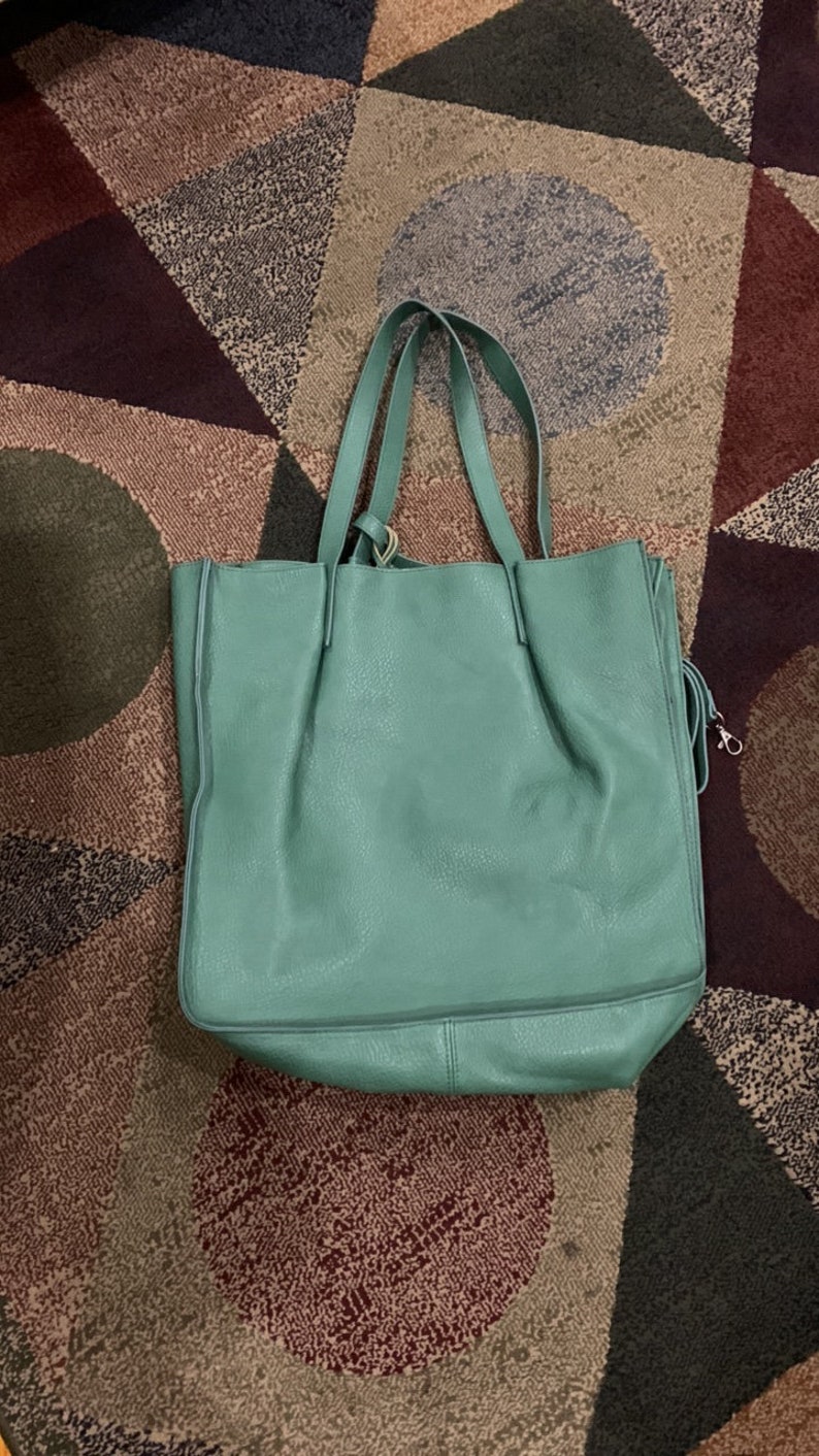 Puede incluir: Un bolso tote grande de cuero turquesa con dos asas. El bolso tiene un aspecto suave y texturizado y una forma rectangular. El bolso est&aacute; sobre una alfombra estampada con varios colores y formas.