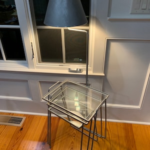 May include: A set of three nesting glass-top tables with a silver metal frame. The tables are arranged in a stack, with the largest table on the bottom and the smallest table on top. The tables are sitting on a wooden floor.