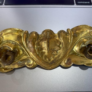 May include: A gold-toned metal decorative hardware piece with ornate floral designs and two attached rings. The piece has a worn, antique look.