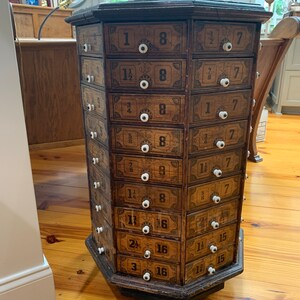 Puede incluir: Antiguo mueble de boticario de madera con m&uacute;ltiples cajones, cada uno etiquetado con n&uacute;meros y fracciones. El mueble tiene pomos blancos y est&aacute; coronado con una escultura de pato de madera. Est&aacute; sobre un suelo de madera.