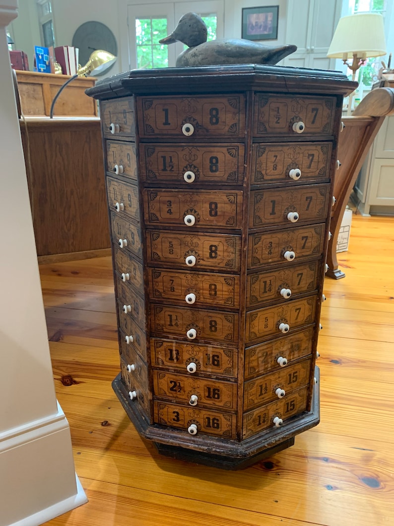 Puede incluir: Antiguo mueble de boticario de madera con m&uacute;ltiples cajones, cada uno etiquetado con n&uacute;meros y fracciones. El mueble tiene pomos blancos y est&aacute; coronado con una escultura de pato de madera. Est&aacute; sobre un suelo de madera.